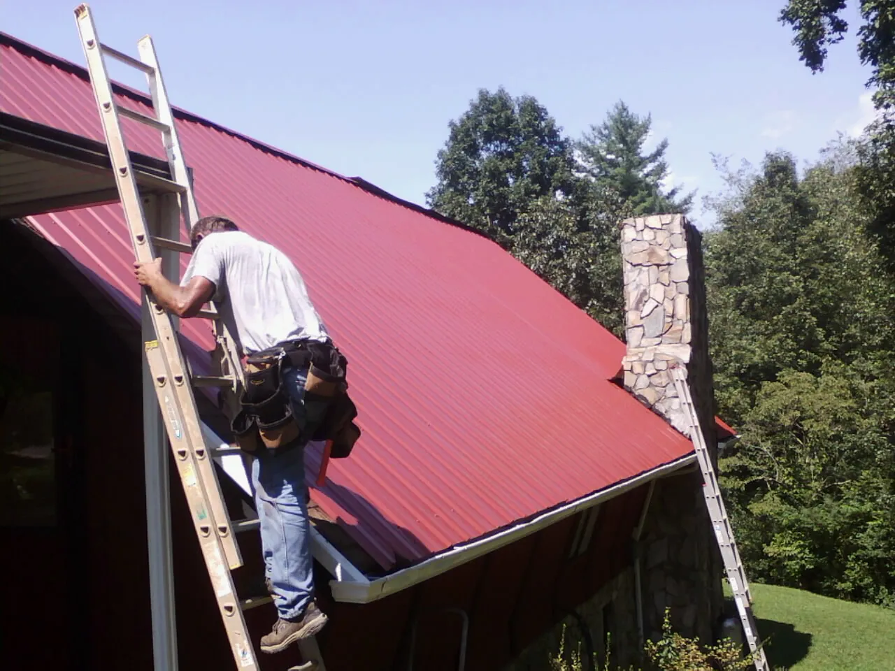 Roofer on a ladder beside a red metal roof and stone chimney — Soffit & Fascia Repair by Roofing Brothers in Lone Tree
